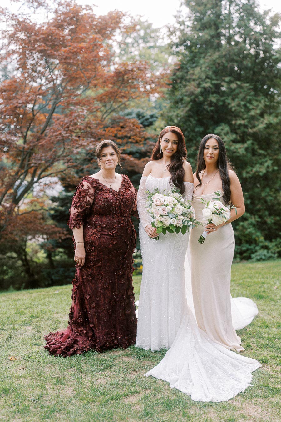 Three women standing on grass in elegant gowns, including a bride in a white wedding dress holding a bouquet of pink and