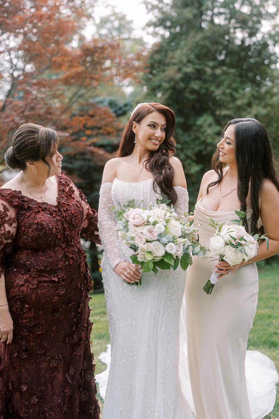 Three women in elegant dresses, holding bouquets, smile outdoors in a lush garden setting. They are surrounded by greenery