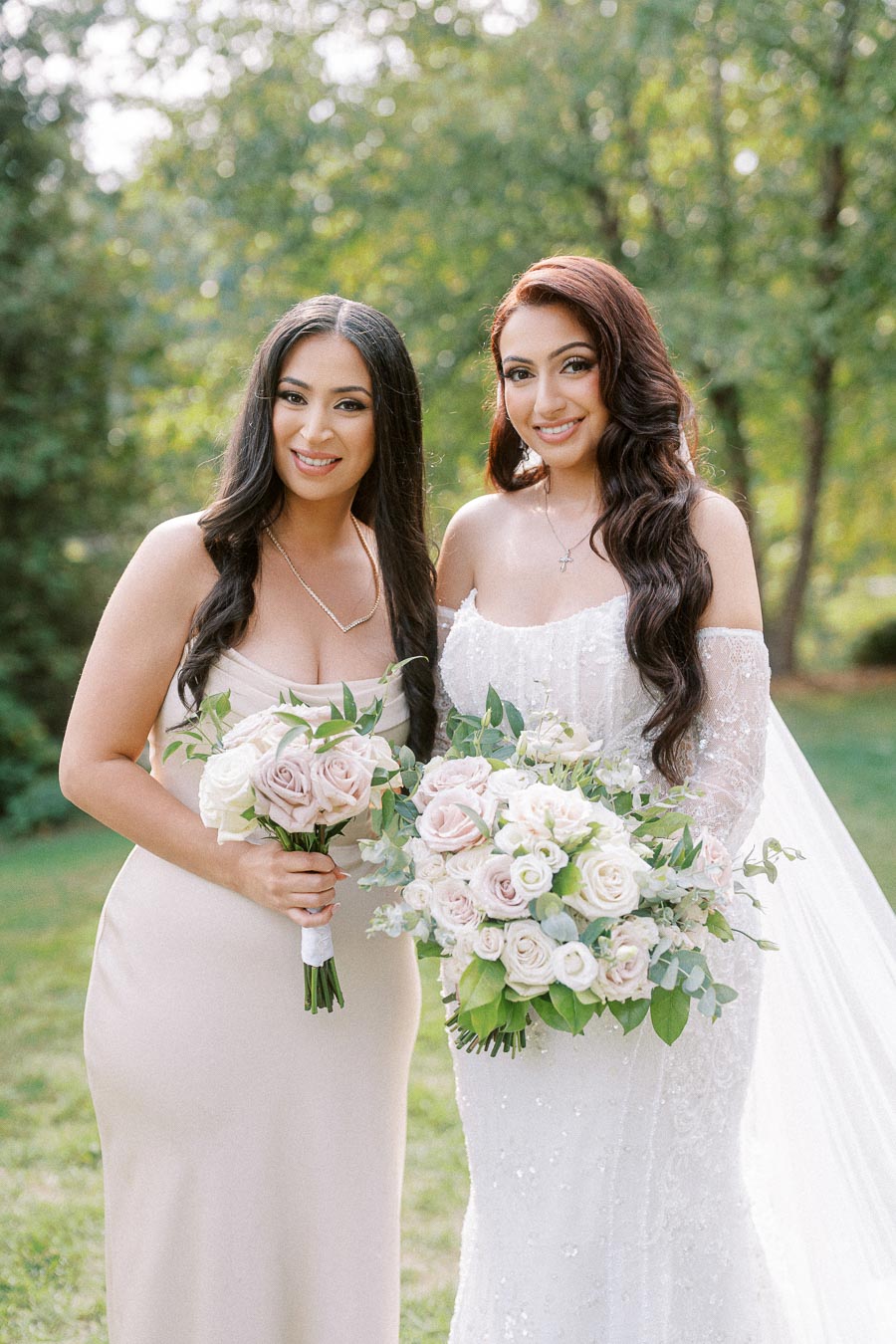 Two women in elegant dresses holding bouquets of pastel roses, standing outdoors in a lush green garden setting.