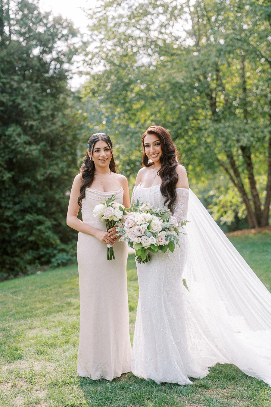 Two women in elegant dresses pose outdoors, holding bouquets of pink and white roses, surrounded by lush greenery in a