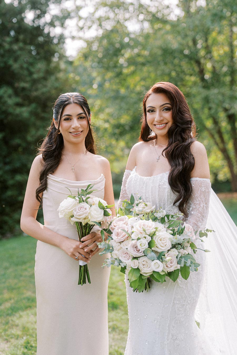 Two women in elegant dresses holding bouquets of pale pink and white roses, standing outdoors in a lush green garden, with