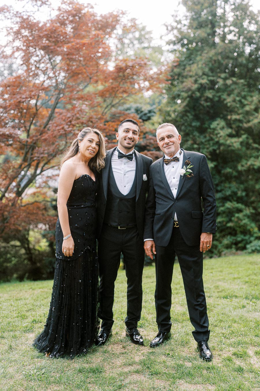 Three people dressed in formal black attire posing outdoors on a grassy area, with vibrant autumn trees in the background.
