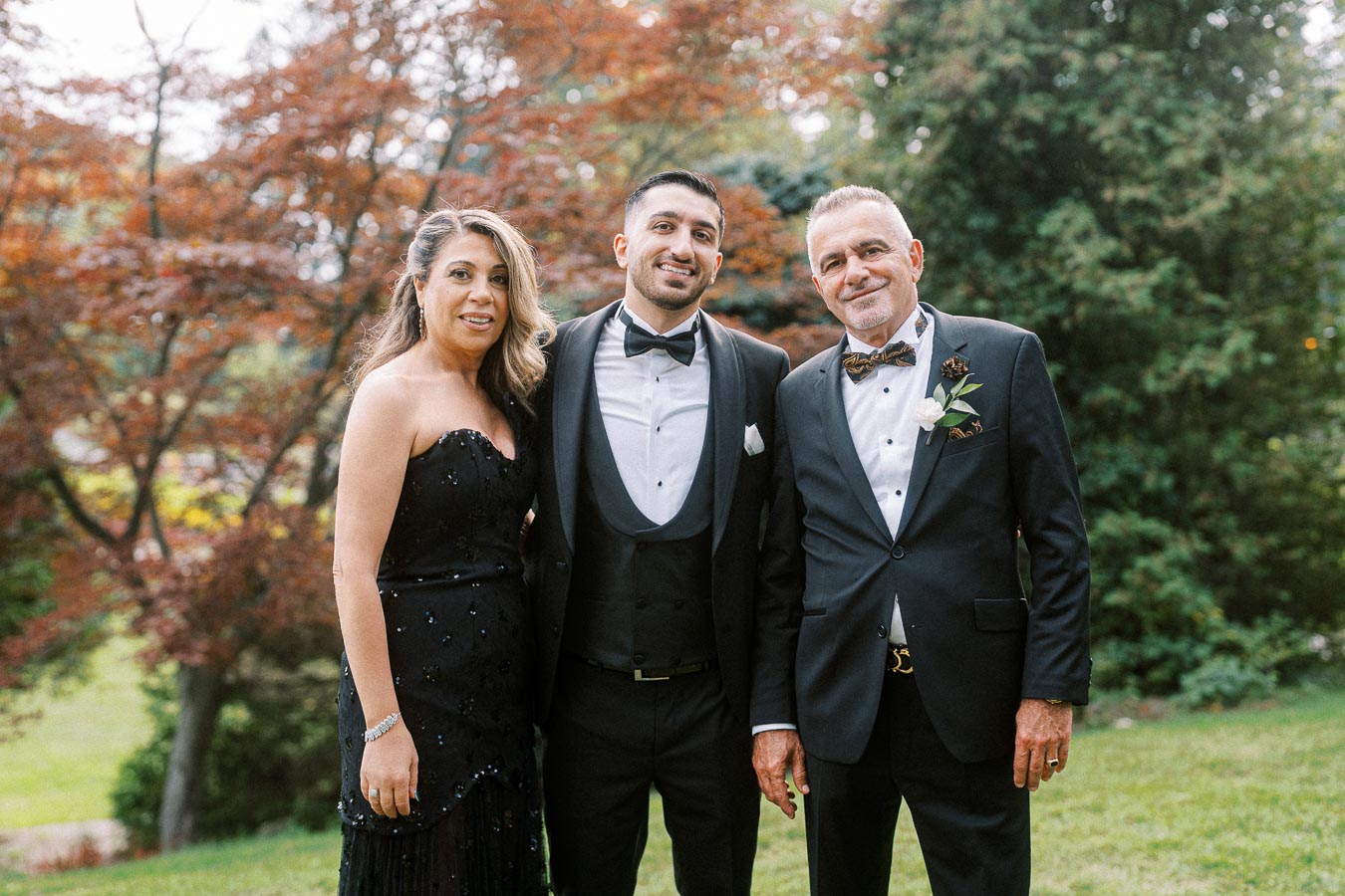 A smiling group of three people dressed in formal attire, standing together outdoors with a background of lush greenery and