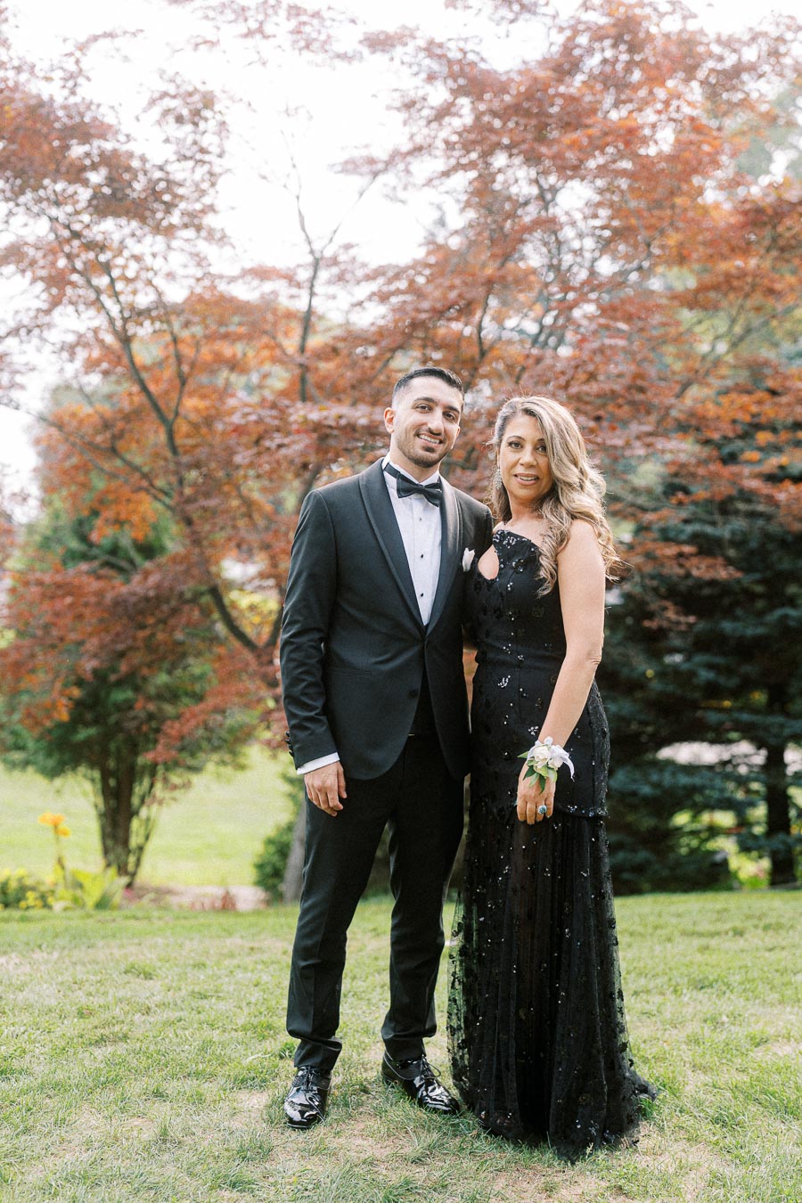 A man in a black tuxedo and a woman in an elegant black gown stand smiling in a lush garden with vibrant trees in the