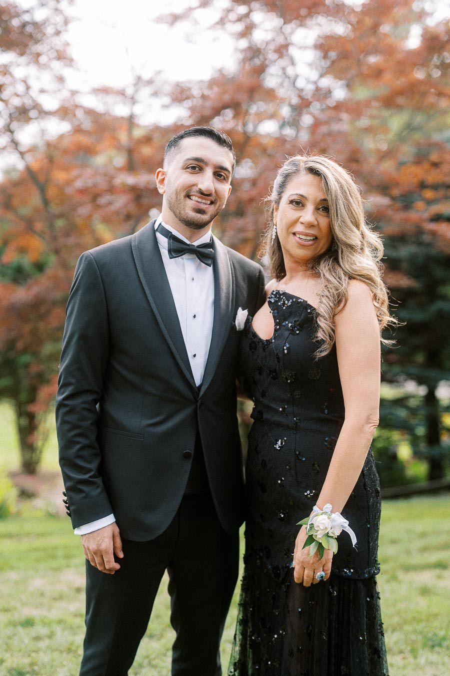A man and woman dressed elegantly pose together outdoors, with the background showcasing lush greenery and autumn-colored