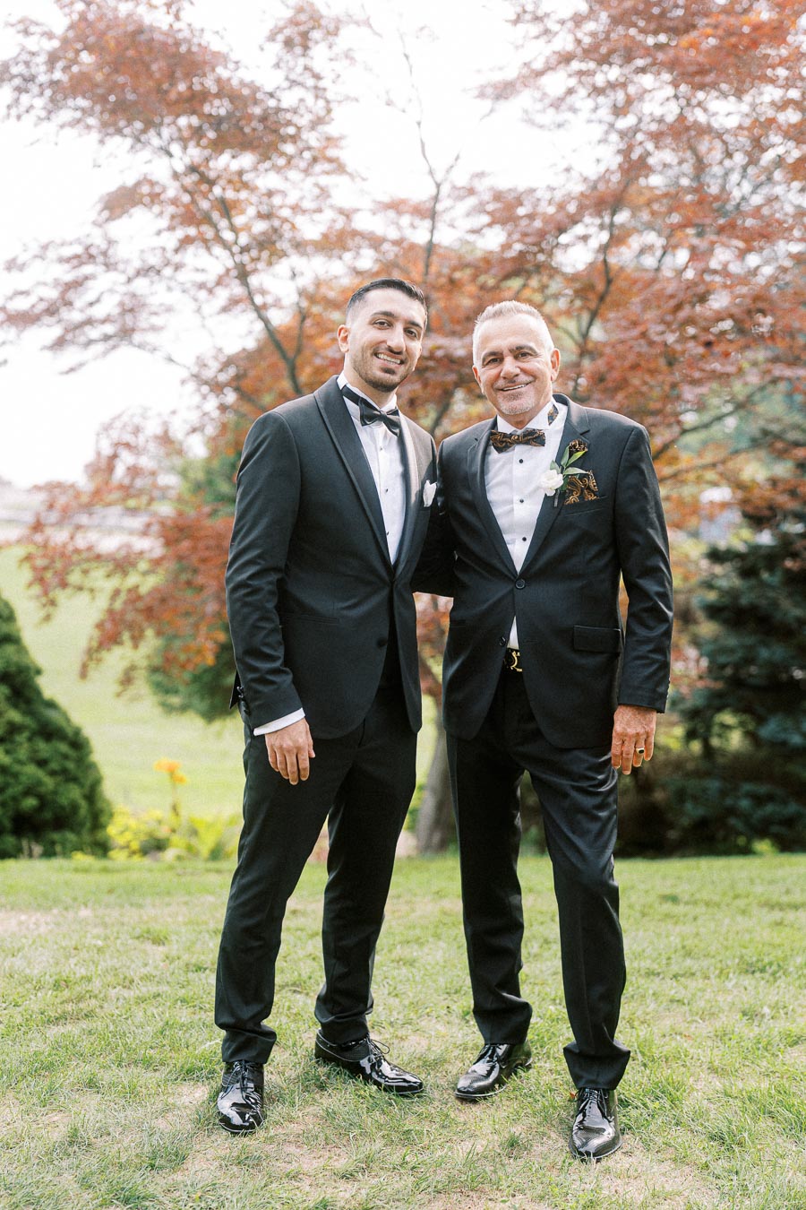 Two men in elegant black tuxedos pose outdoors, surrounded by lush greenery and vibrant foliage, showcasing a stylish