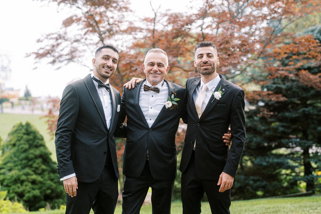 A group of three men in elegant suits smiling and posing together outdoors, surrounded by lush greenery and trees in the