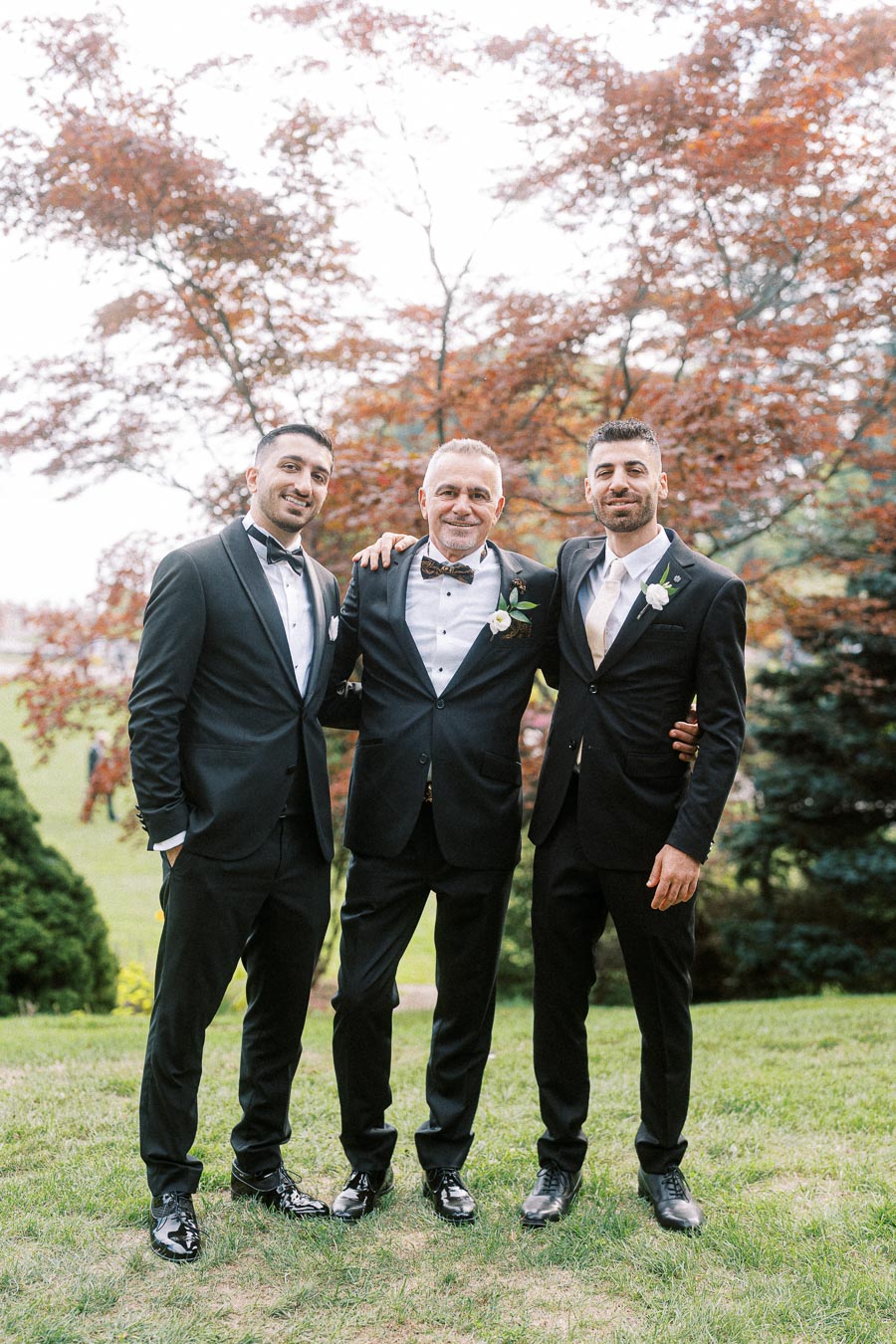A group of three men in elegant black tuxedos with white shirts and bow ties, standing together on grass in front of