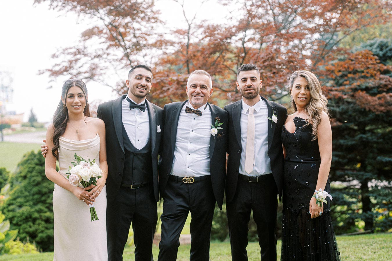 A group of five people, dressed in formal attire, pose together outdoors in front of vibrant green and reddish foliage, each