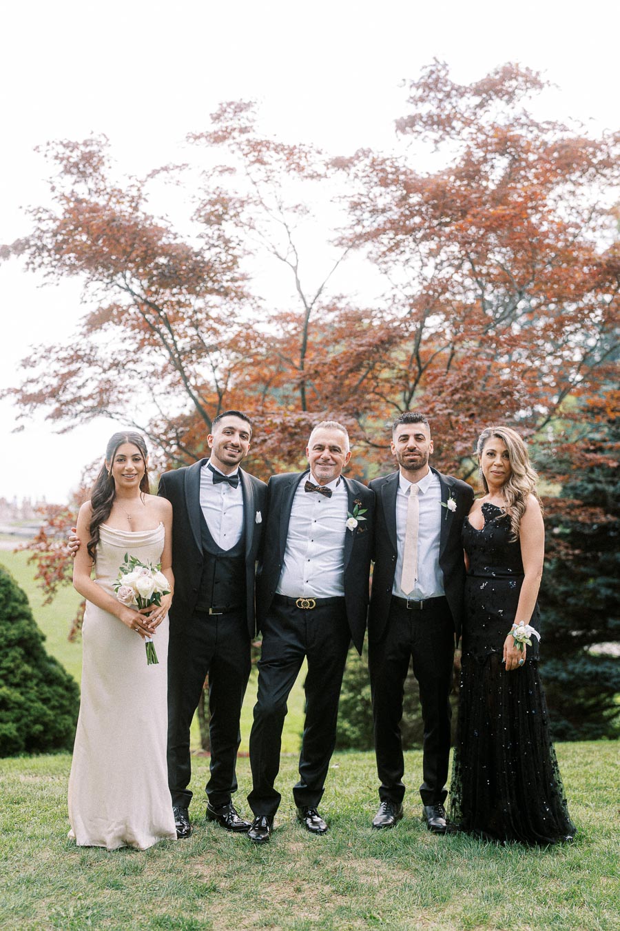 A family poses elegantly in formal attire outdoors, with a green lawn and red-leaved tree in the background, capturing a