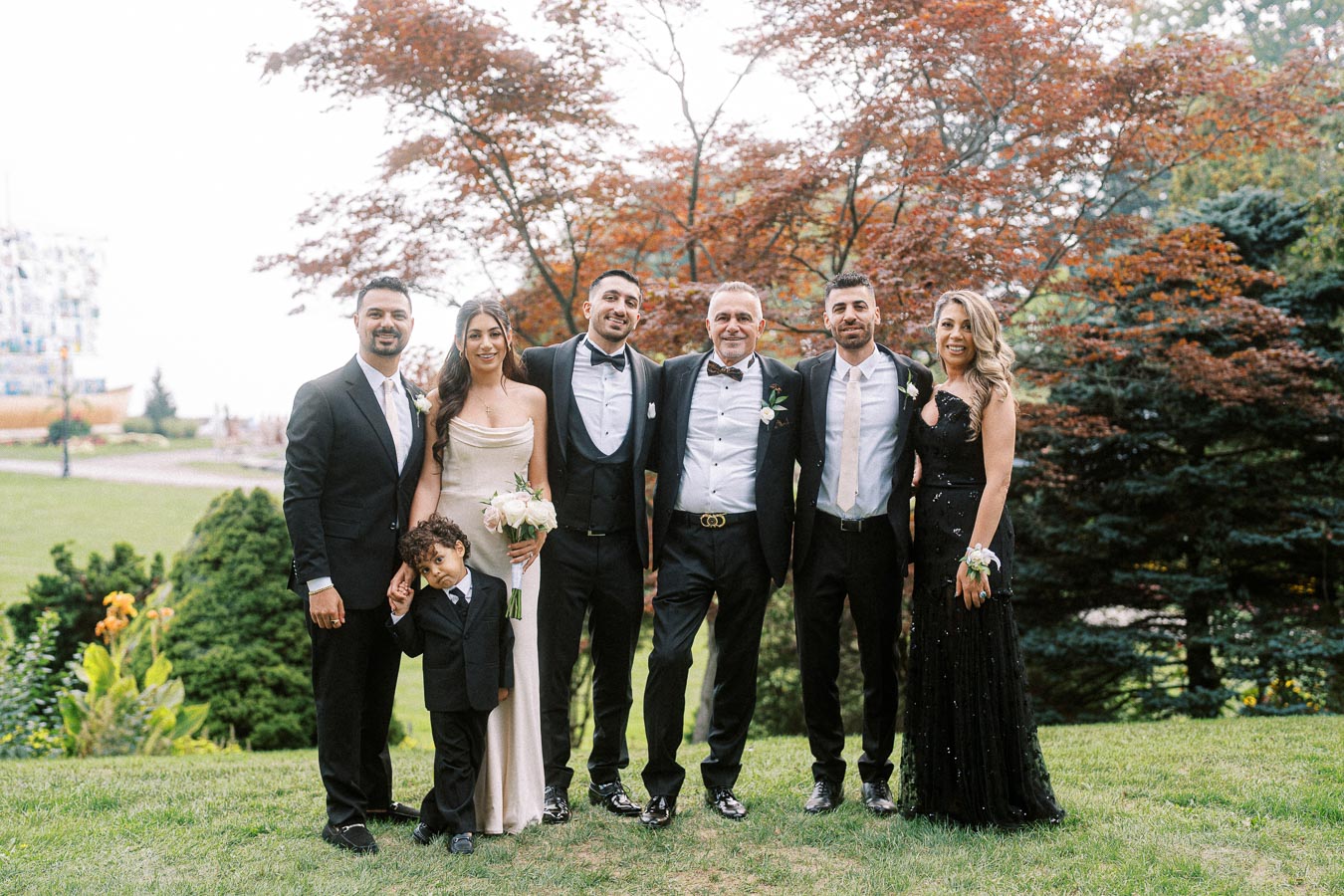 A group of seven people dressed in formal attire standing on a lush green lawn with trees in the background, posing and