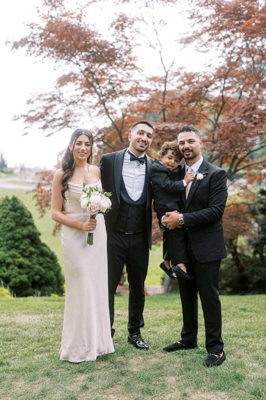 Group of people dressed in formal attire posing outdoors with a garden backdrop; two men in suits, a woman in an elegant