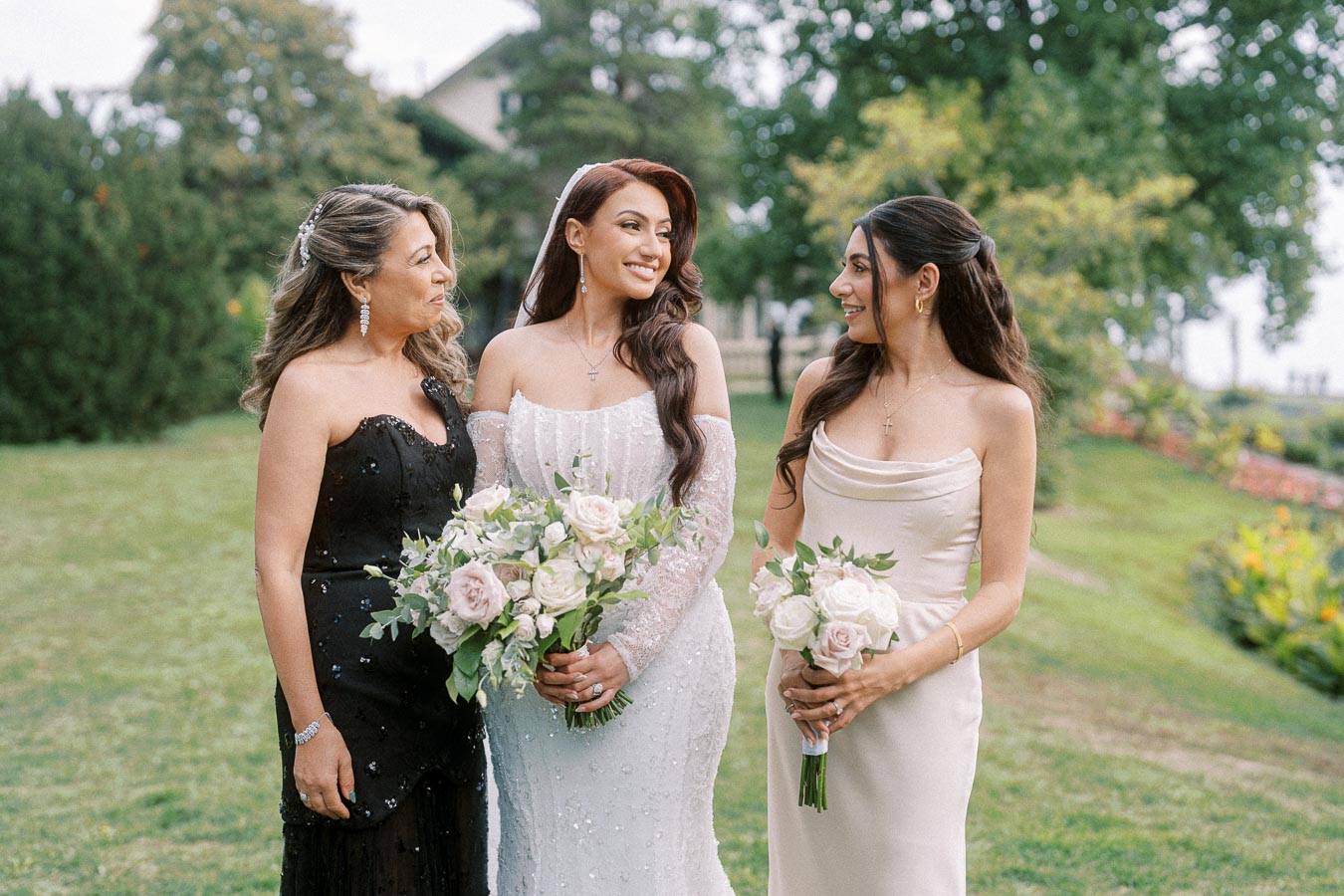 Three women in elegant dresses, standing together in a lush garden setting, smiling and holding bouquets of white and pink