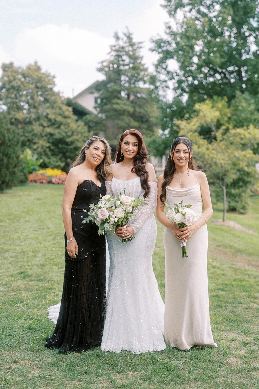 Three women in elegant dresses stand together outdoors on a grassy lawn. One woman in a white gown holds a bouquet of light