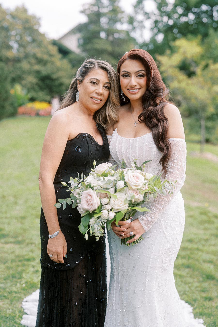 Two women at an outdoor wedding smiling; the bride in a white lace gown holding a bouquet of roses and greenery, and a woman