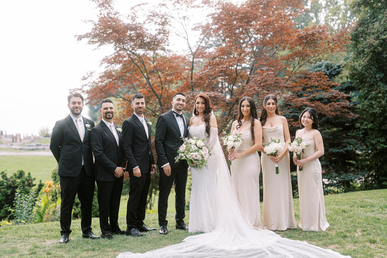 Wedding party posing outdoors with elegant attire, featuring a bride in a white gown holding a bouquet, surrounded by