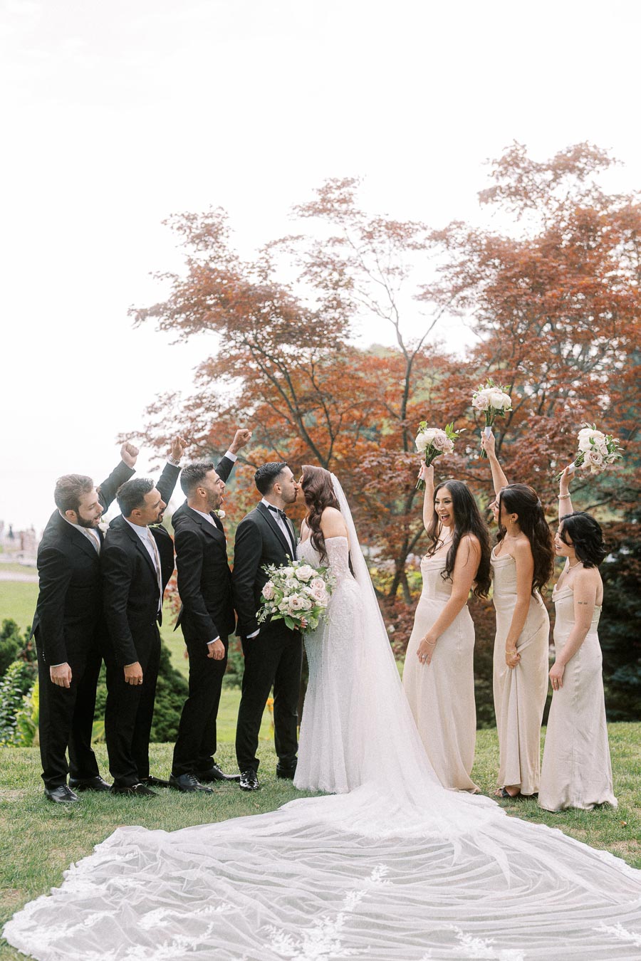 Bride and groom kissing outdoors with bridesmaids and groomsmen celebrating, surrounded by lush greenery and blooming trees.