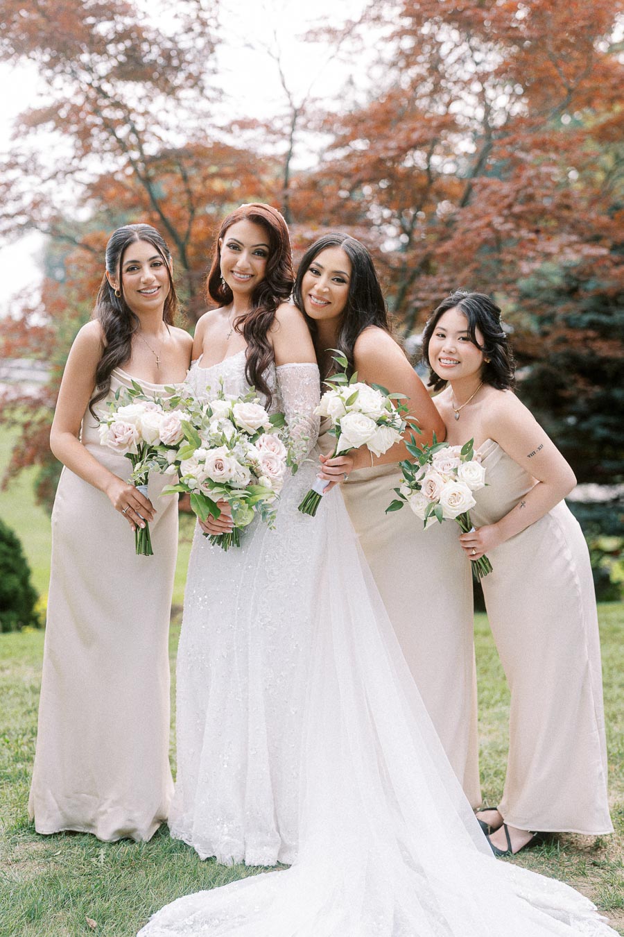 Elegant bridal party posing outdoors with the bride in a white lace gown surrounded by bridesmaids in beige dresses, holding
