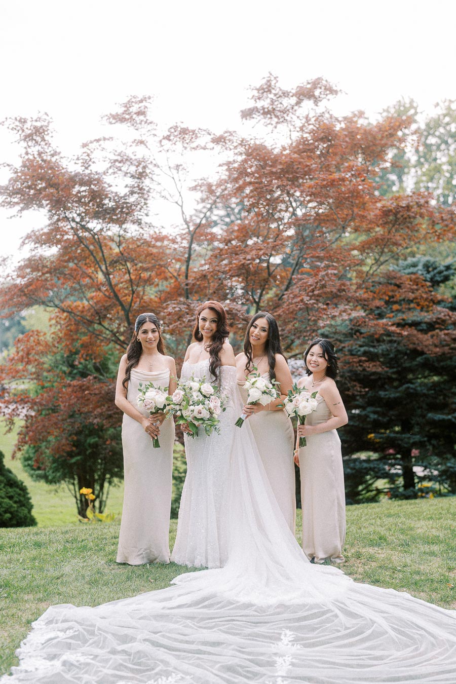 Beautiful bride with three bridesmaids in pale dresses holding bouquets, posing outdoors with lush greenery and trees in the