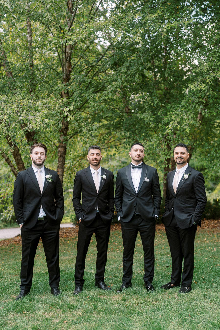 Four groomsmen in black suits and white dress shirts standing together on a lush green lawn with trees in the background,