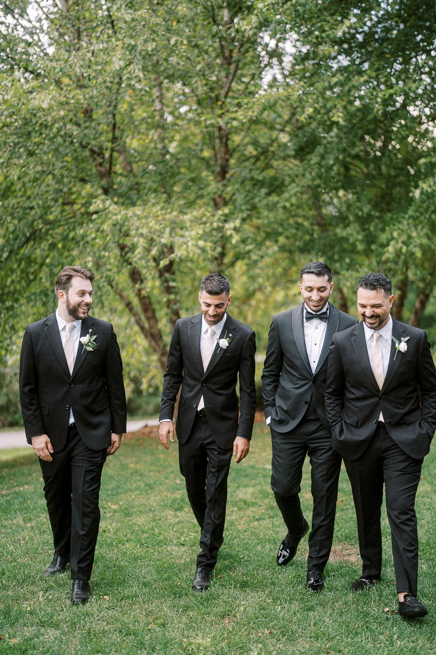 Four groomsmen in black suits walking together on a lush green lawn, surrounded by trees, enjoying a moment of camaraderie