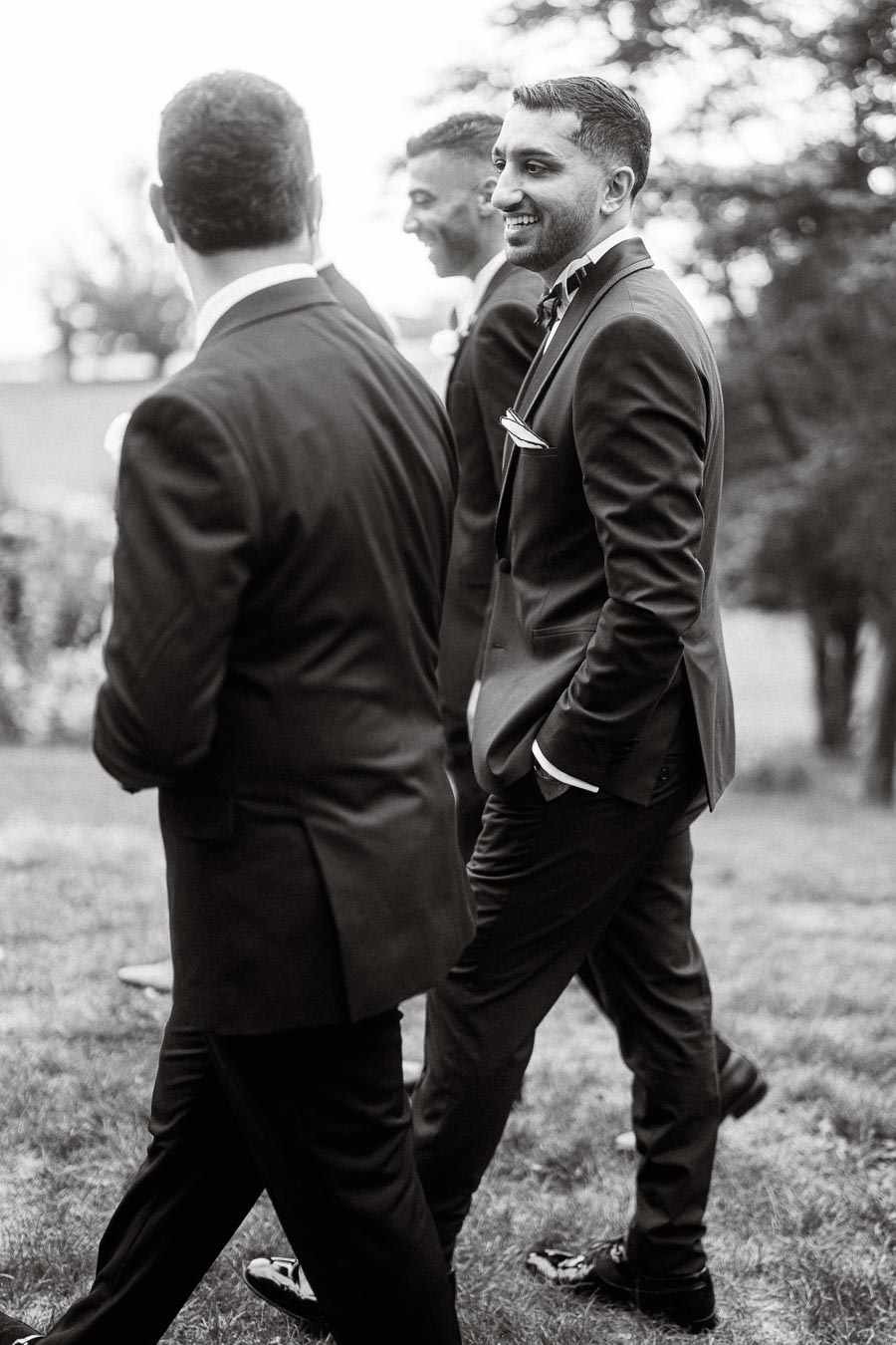 Black and white photo of three men in suits walking outdoors, smiling and engaged in conversation, captured in a candid