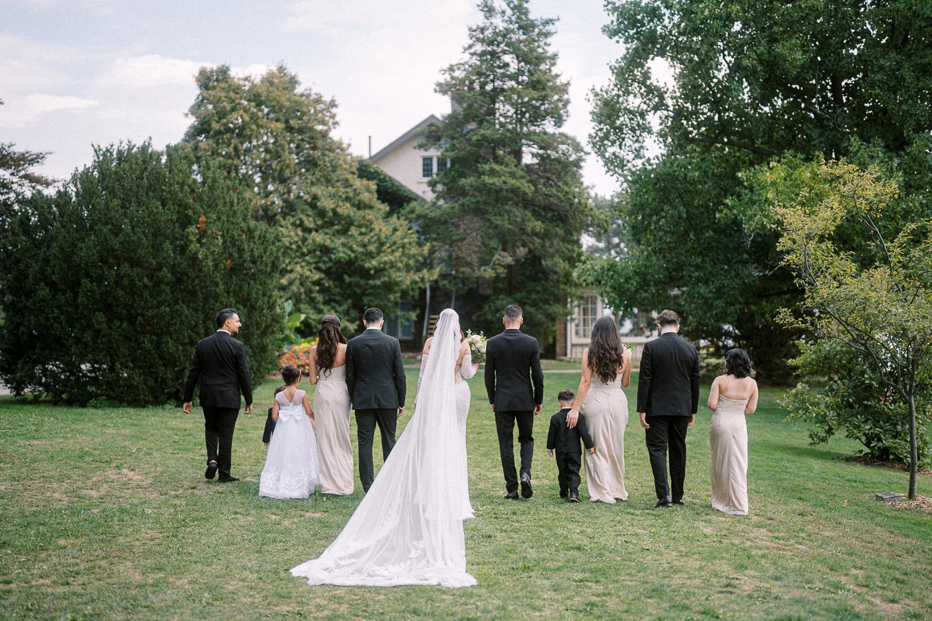 A wedding party walks across a lush green lawn towards a scenic garden, with bridesmaids in elegant dresses and groomsmen in