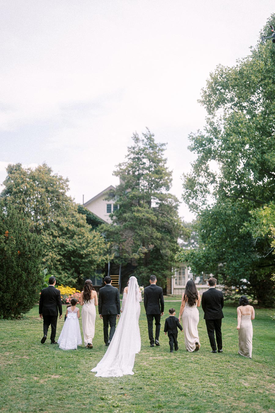 A wedding party, including a bride in a flowing white gown and veil, walks towards a house surrounded by lush greenery and