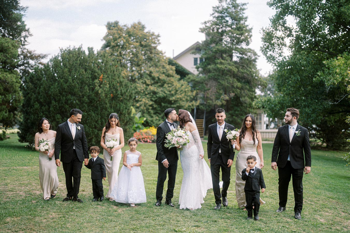 Wedding party walking together on a grassy lawn with the bride and groom kissing in the center, surrounded by bridesmaids in