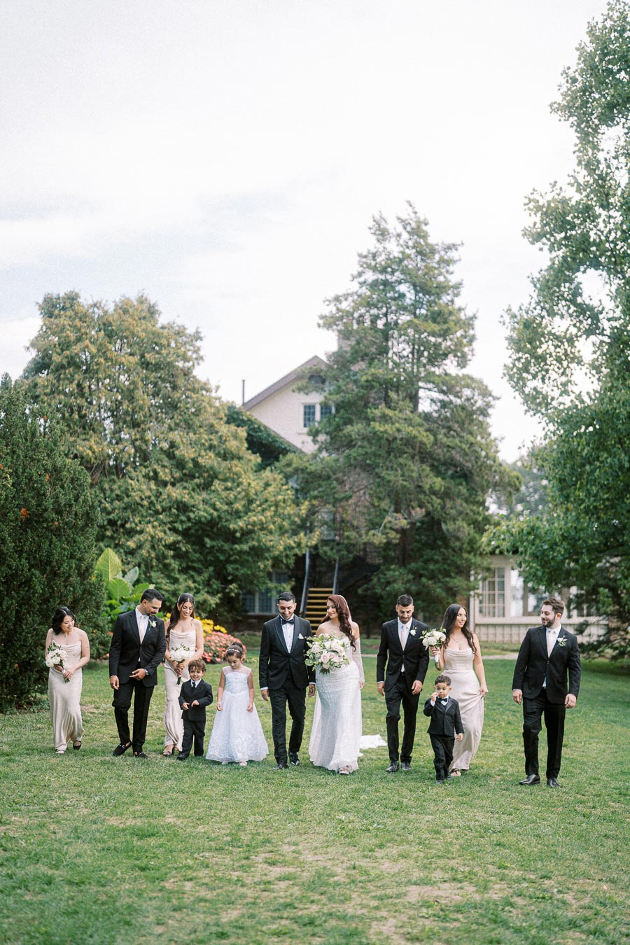 A wedding party walks outside on a grassy lawn, with trees and a house in the background. The bride, groom, bridesmaids,