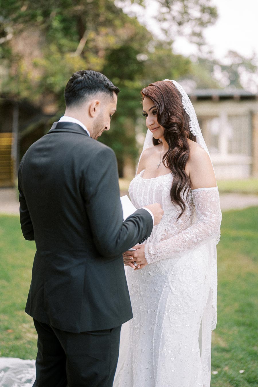 A bride in a white lace wedding gown listens to her groom reading vows during an outdoor ceremony, surrounded by lush