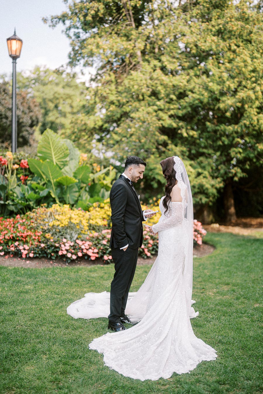 A couple on their wedding day in a garden setting, with the bride in a white lace gown and veil, and the groom in a black