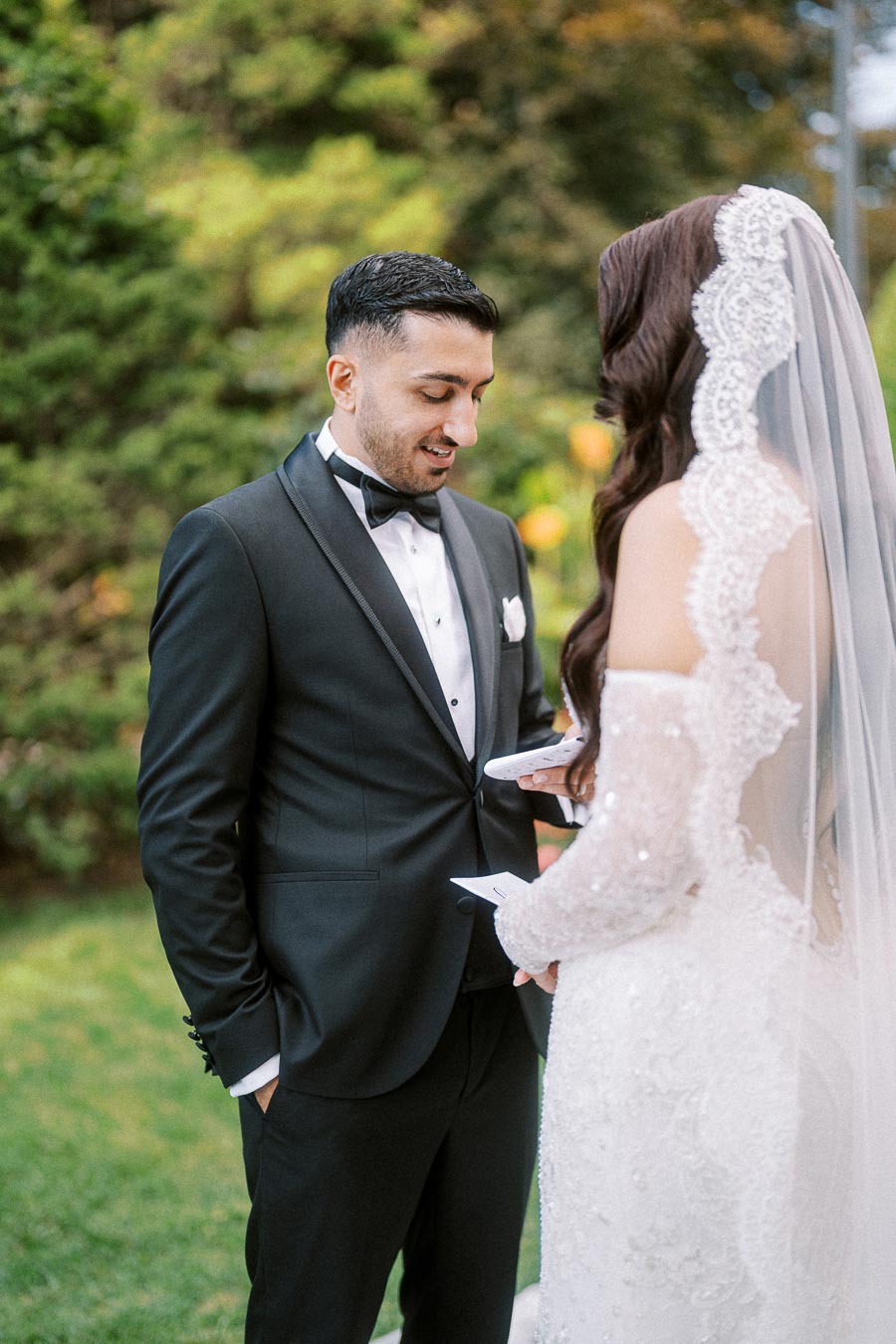 A bride and groom exchanging vows outdoors on their wedding day, with the groom in a black tuxedo and the bride in a white