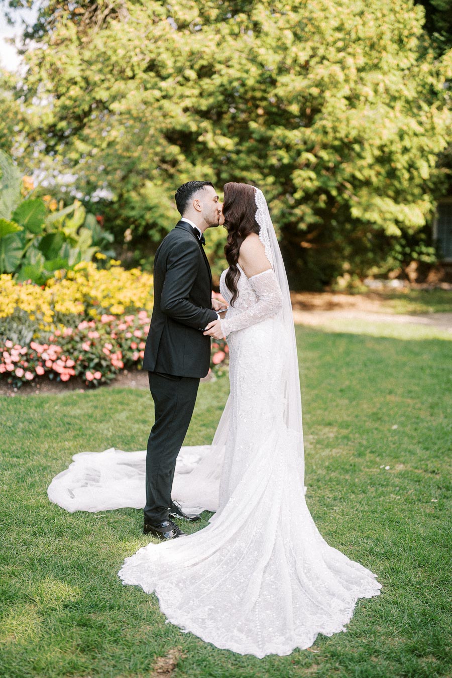 A bride and groom share a kiss in a lush garden, with the bride wearing a lace wedding gown and veil, and the groom in a