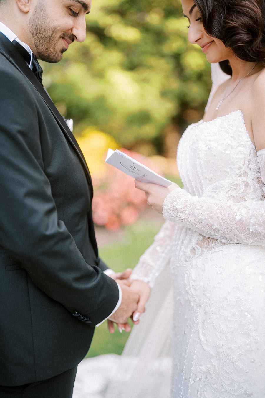 A bride and groom holding hands during their wedding ceremony, with the bride reading vows from a booklet. The scene is set