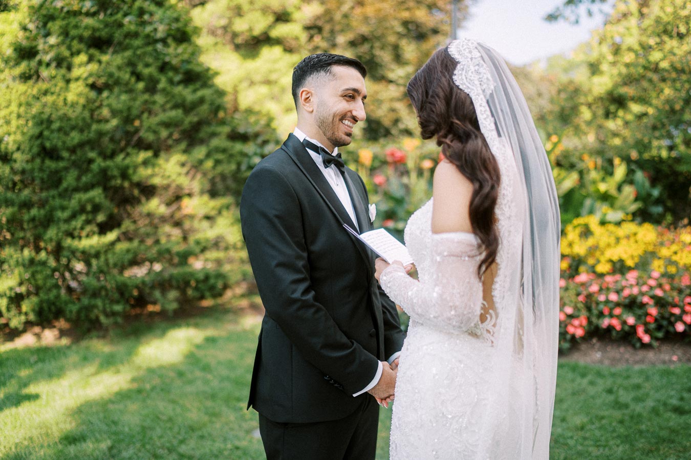 Bride and groom exchanging vows outdoors in a garden setting, with colorful flowers and greenery in the background,