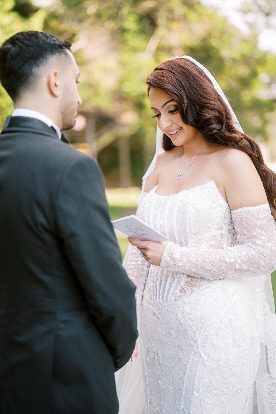 A bride in an elegant white lace wedding dress reads vows to the groom during an outdoor ceremony, surrounded by lush