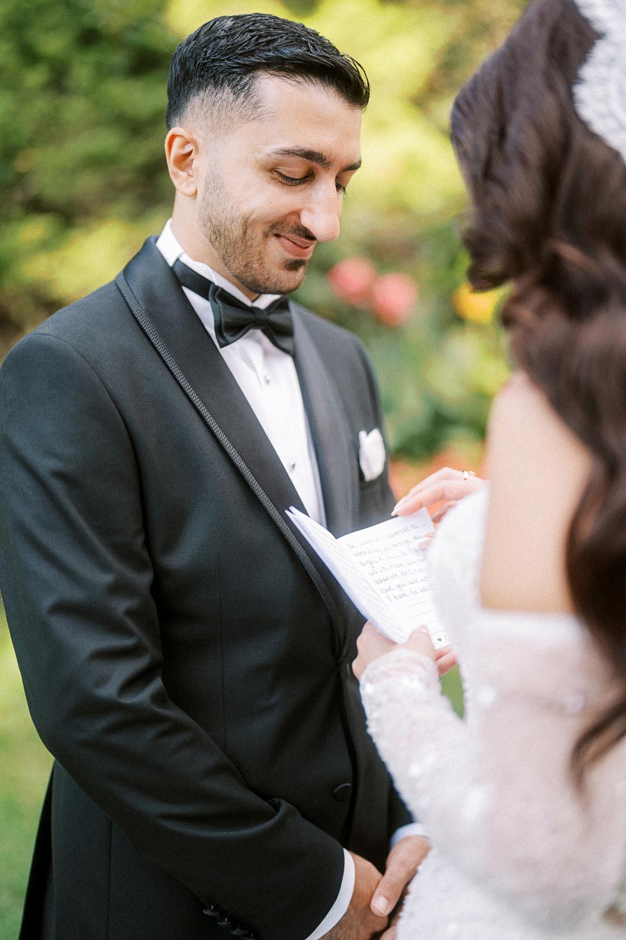 Groom in black tuxedo smiles as he listens to bride reading vows in an outdoor wedding.