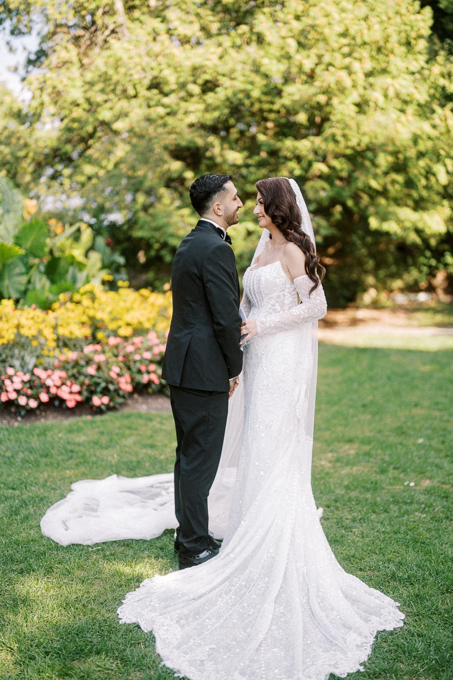 Bride and groom smiling at each other during outdoor wedding ceremony, surrounded by lush greenery and vibrant flowers.
