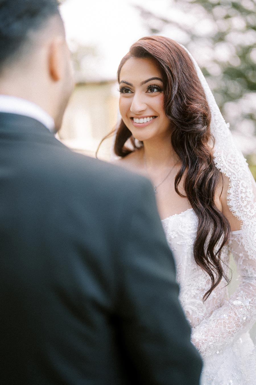 A bride with long wavy hair smiles joyfully during a wedding ceremony, wearing a lace veil and white gown, as she looks at