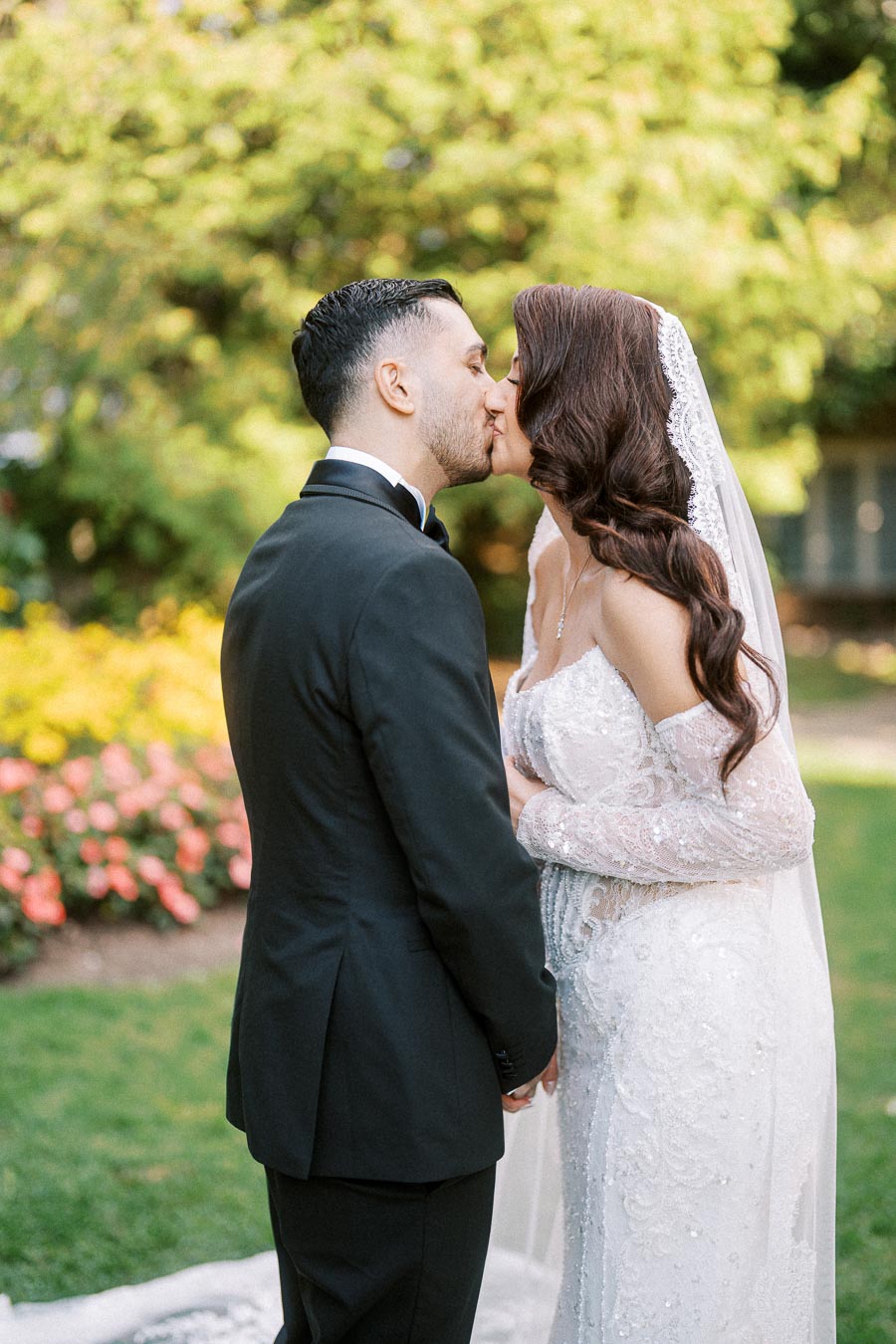 A bride and groom sharing a kiss in a lush garden setting. The bride is wearing an elegant lace wedding dress and veil,
