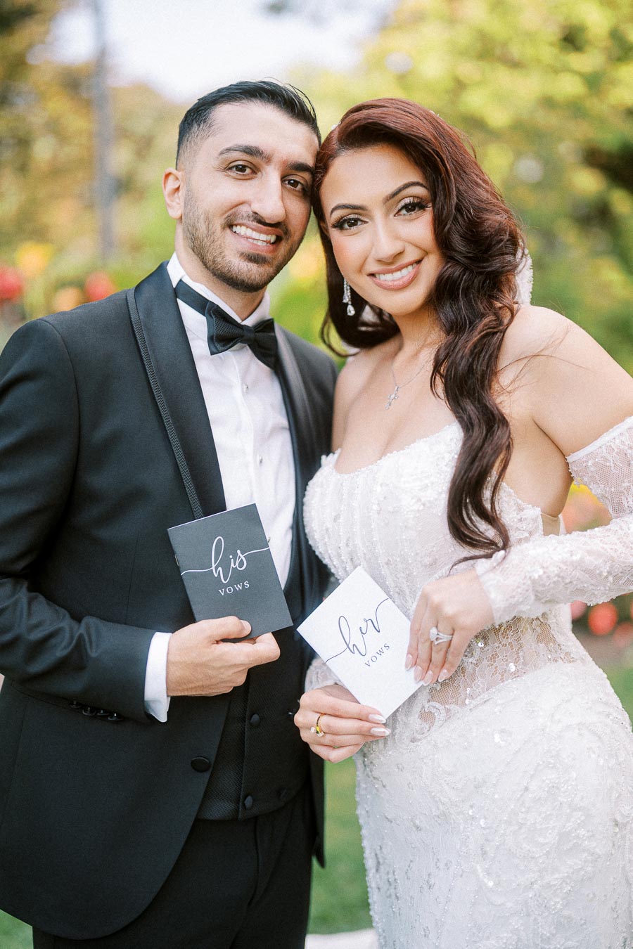 A happy couple in wedding attire holding his and her vow booklets, smiling outdoors in a garden setting.