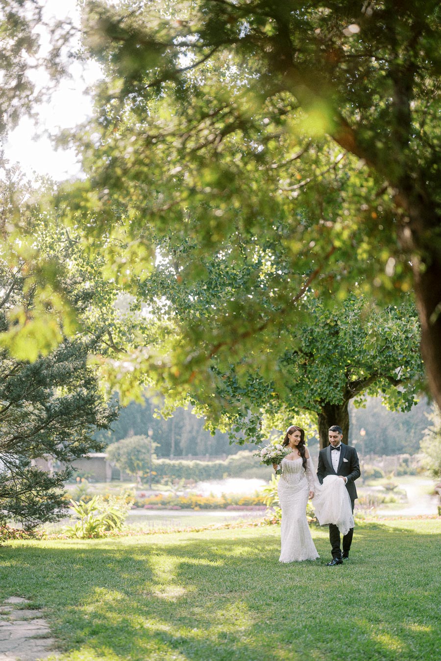 A bride and groom walk hand in hand under a lush green tree, the bride holding a bouquet of flowers. The couple is elegantly