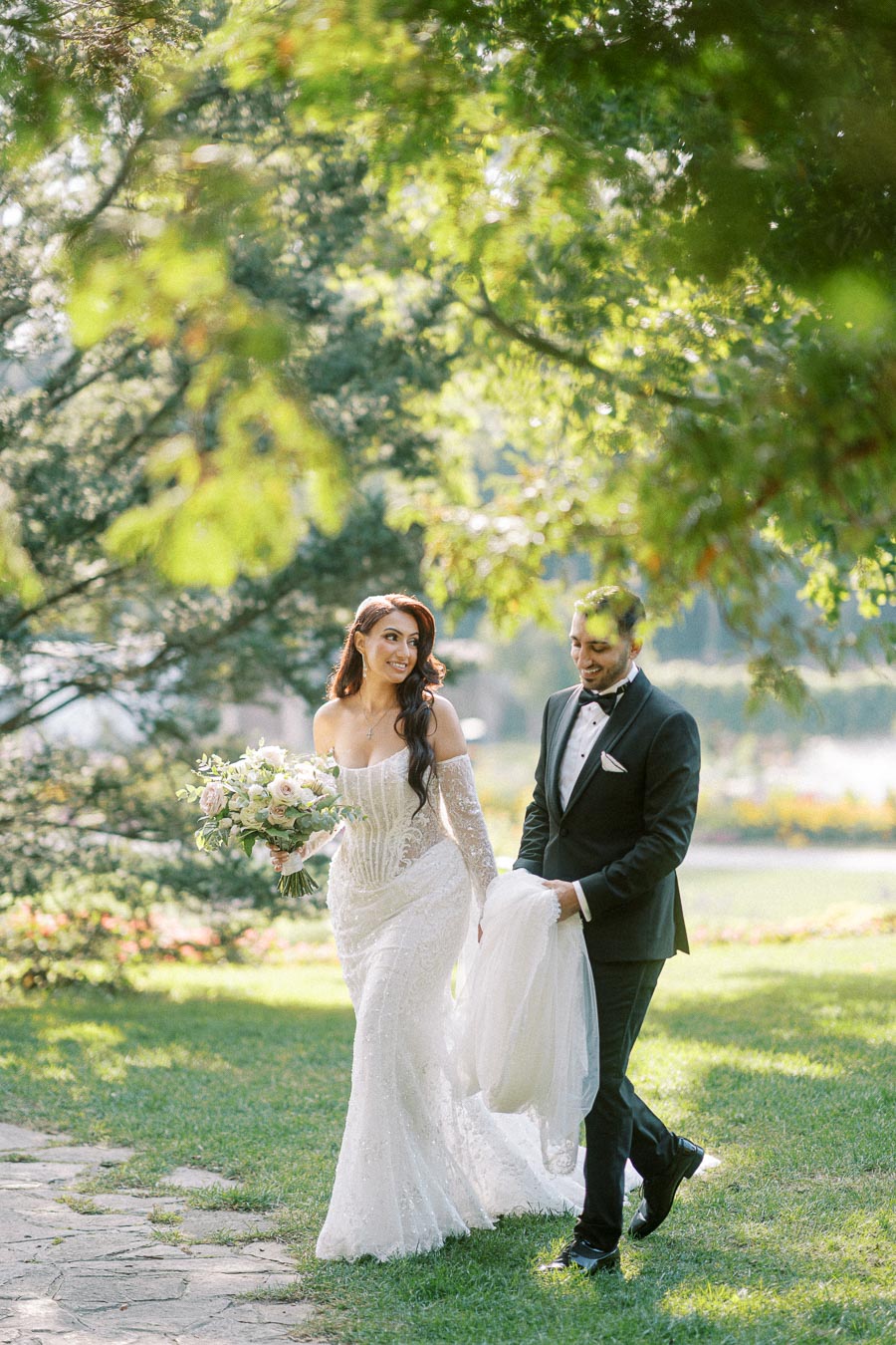 A bride and groom joyfully walk hand in hand through a sunlit garden, with the bride in a stunning white wedding dress