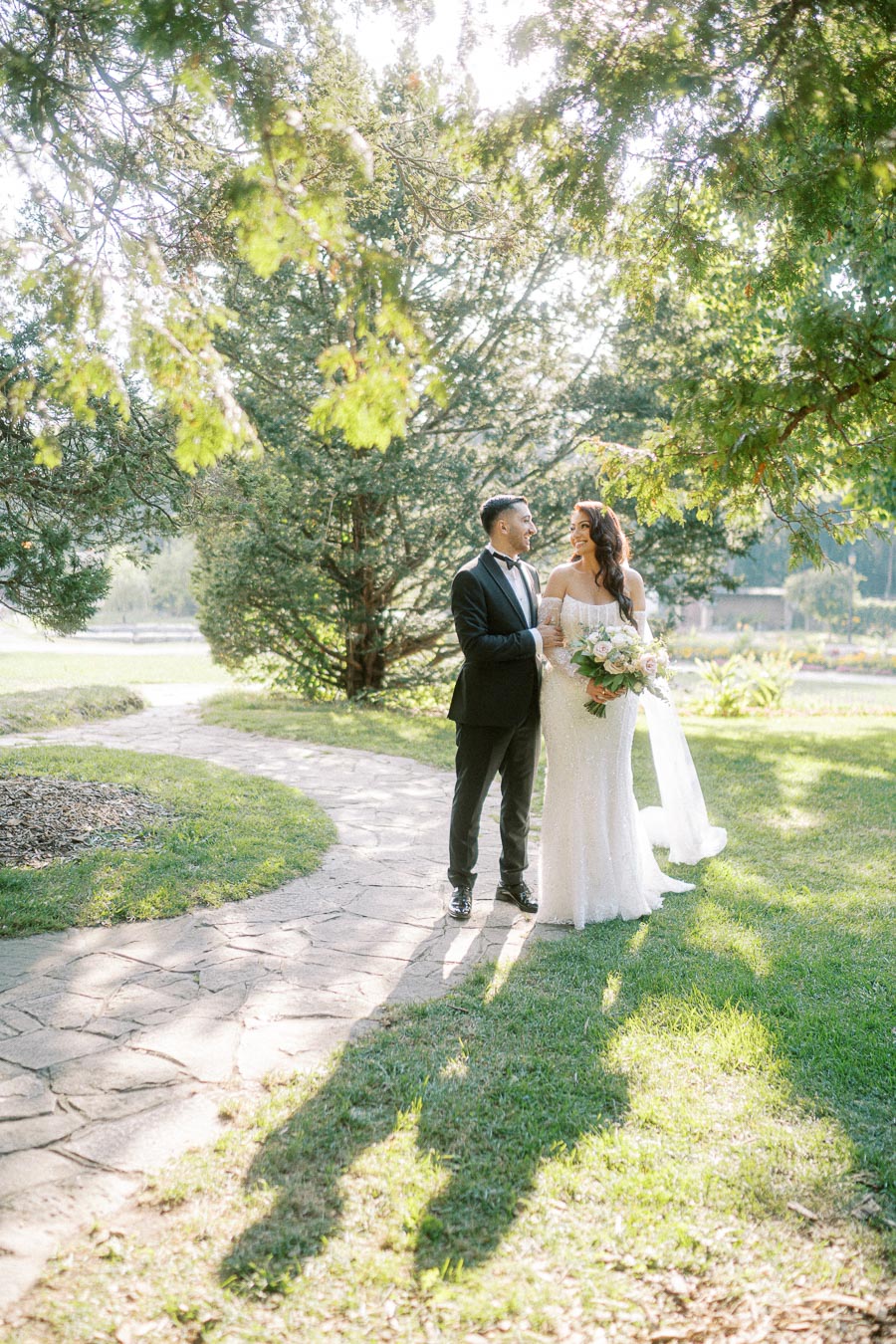 A bride and groom standing together on a sunlit garden path, surrounded by lush greenery and tall trees, capturing a