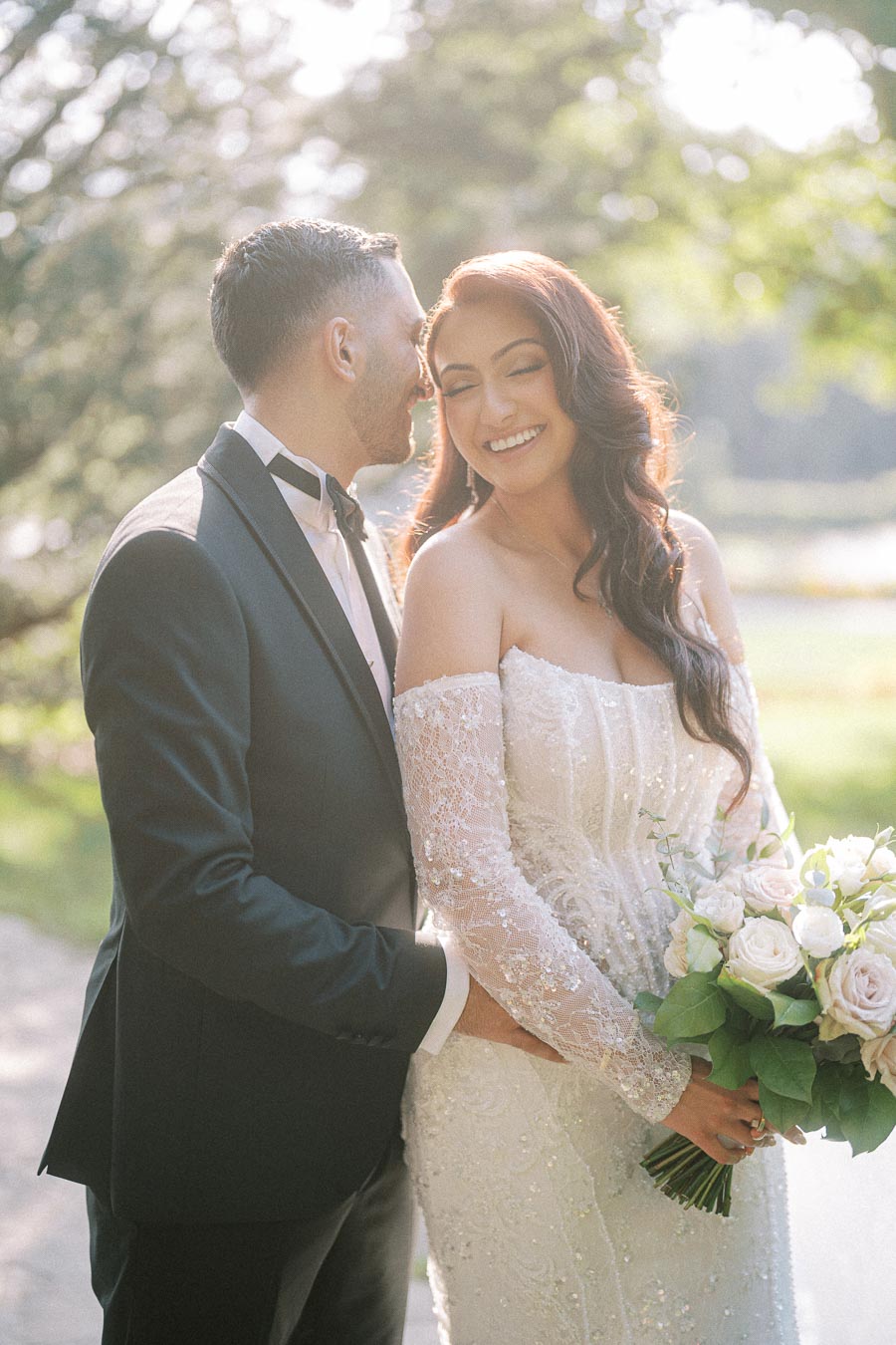 A happy couple on their wedding day, the bride in an elegant off-shoulder lace gown holding a bouquet of white roses, and