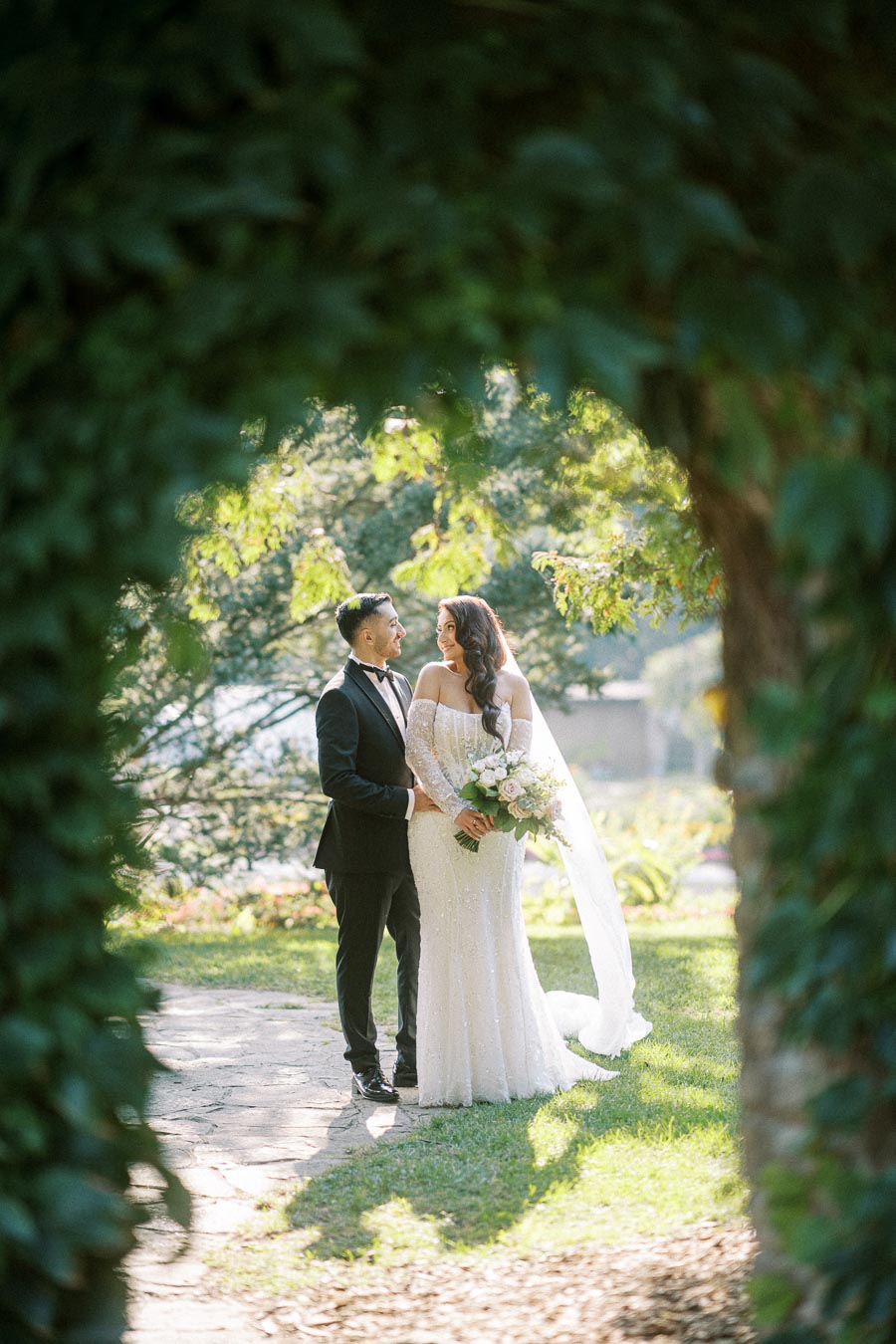 Bride and groom standing together in a lush garden, the bride holding a bouquet of white and pink flowers, surrounded by