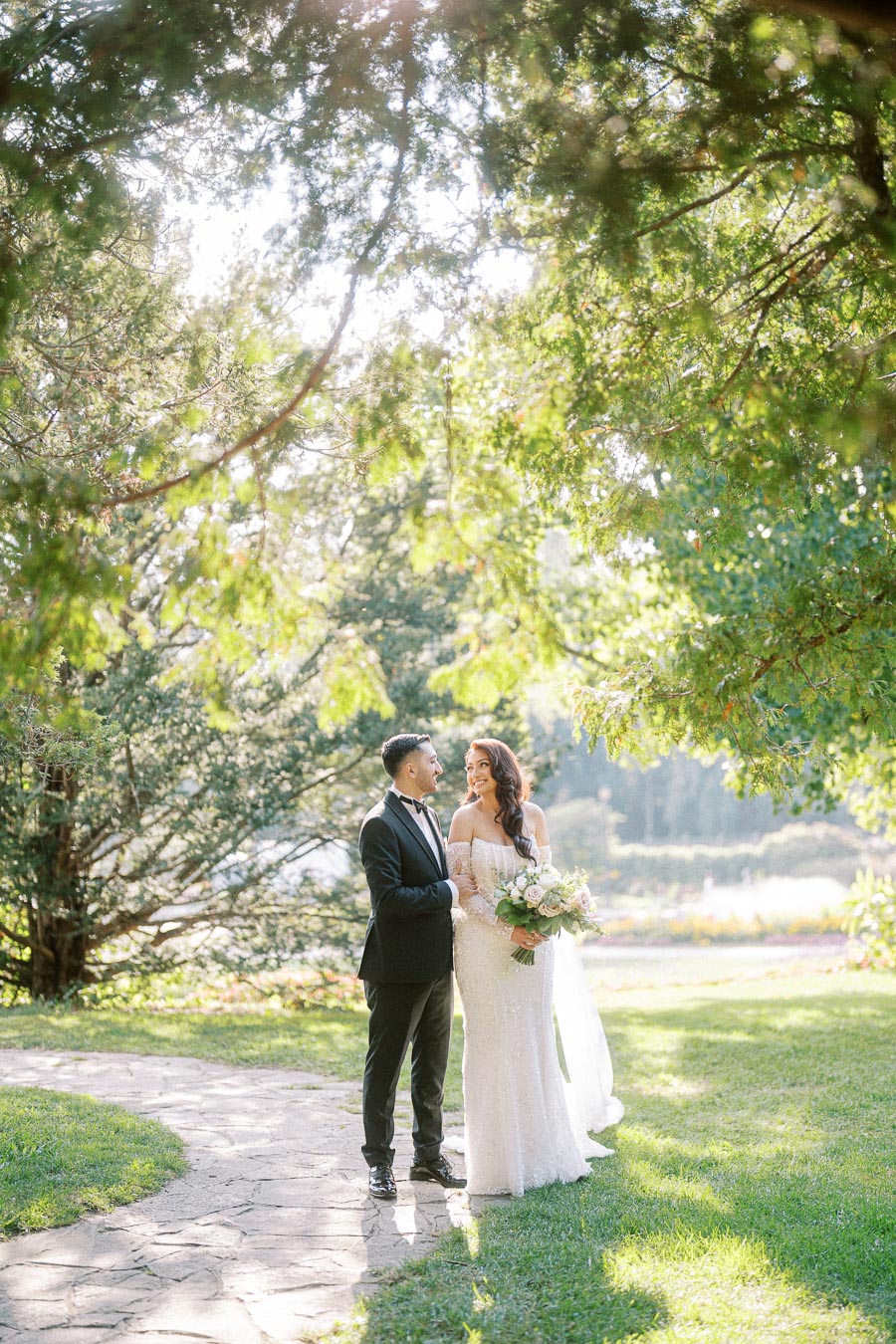 A bride and groom stand together in a sunlit garden, surrounded by lush green trees. The bride is wearing a white dress and