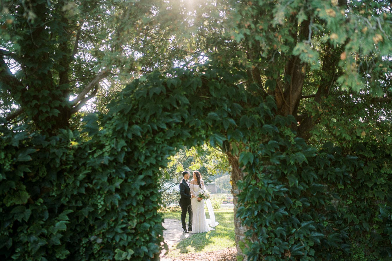 A bride and groom stand together, framed by lush greenery and sunlight filtering through trees, creating a picturesque