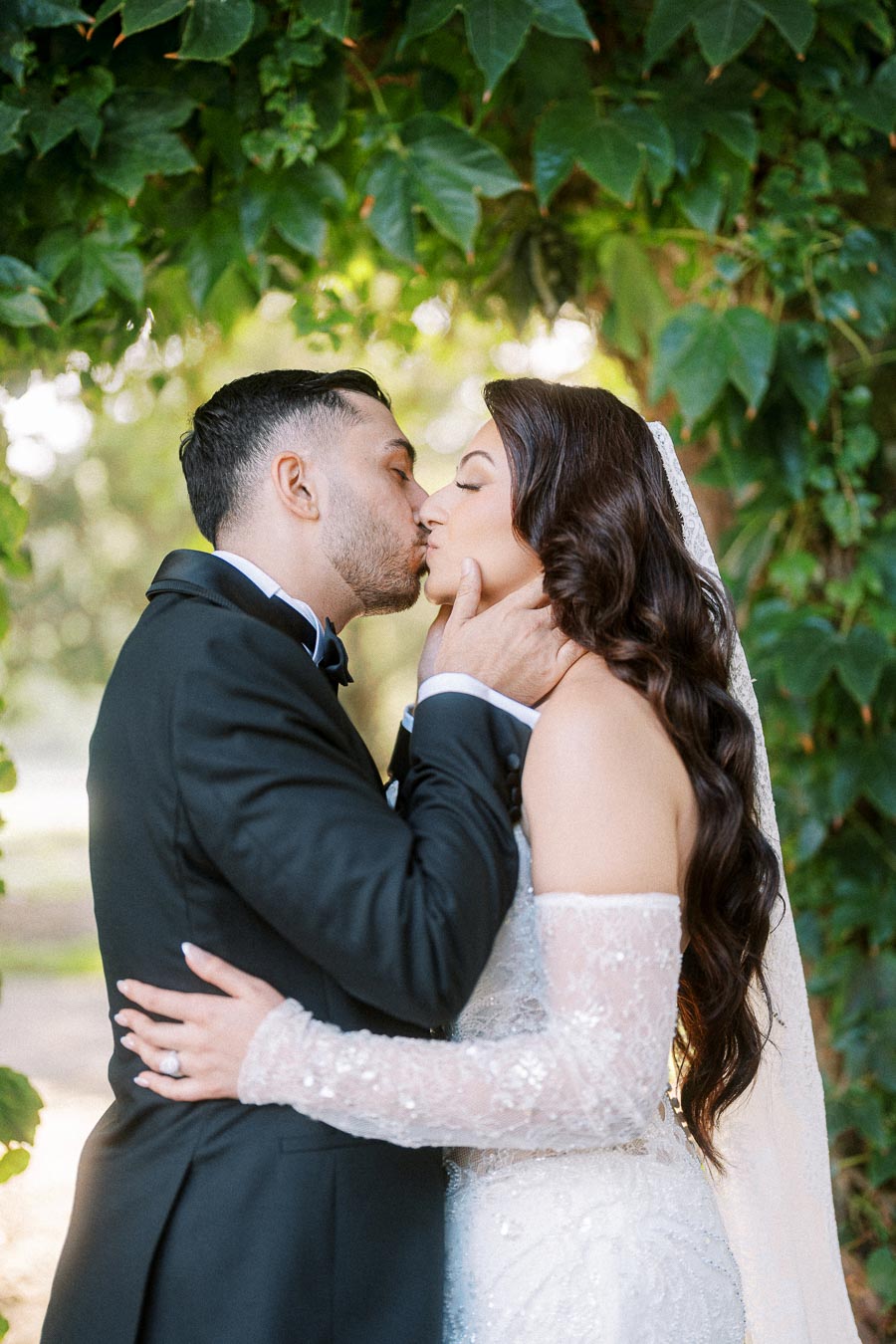 Romantic wedding couple kissing under lush green foliage, bride in elegant off-shoulder lace gown and groom in classic black